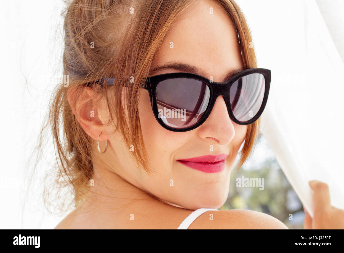 Nahaufnahme der lächelnde junge Frau mit Sonnenbrille befasst sich zurück, während weiße Vorhänge umklammert betrachtet mit Hintergrundbeleuchtung Stockfoto
