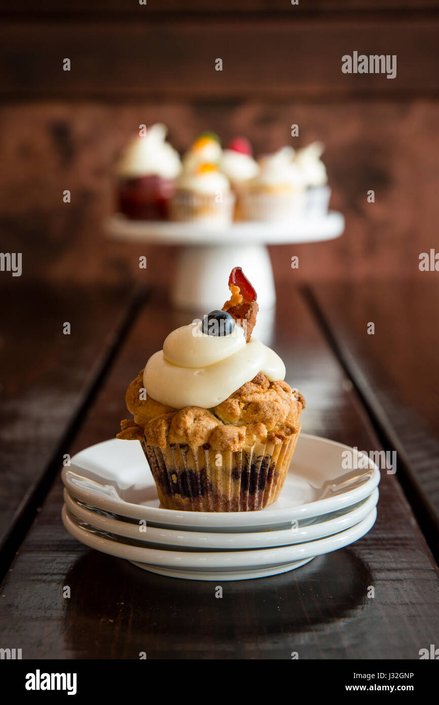 Nahaufnahme von einem Speck Heidelbeere Frühstück Cupcake auf einen Stapel von weißen Platten mit einem Display von Gourmet-Cupcakes im Hintergrund Stockfoto