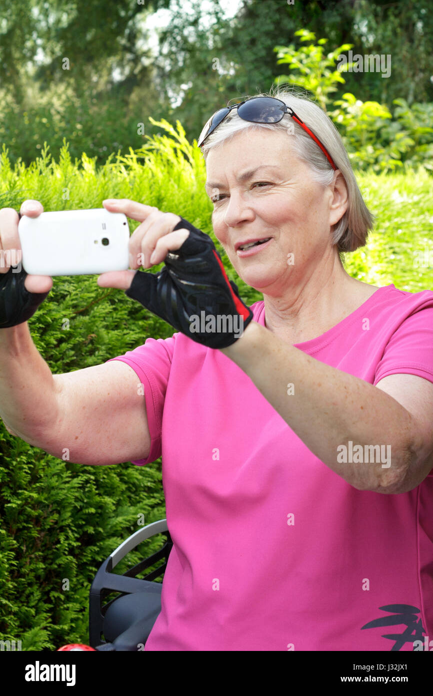 Ältere Frau im Freien im Sommer mit rosa Sport Shirt, Fahrrad Handschuhe und Sonnenbrille ein Foto mit Ihrem Smartphone, grüner Hintergrund Stockfoto