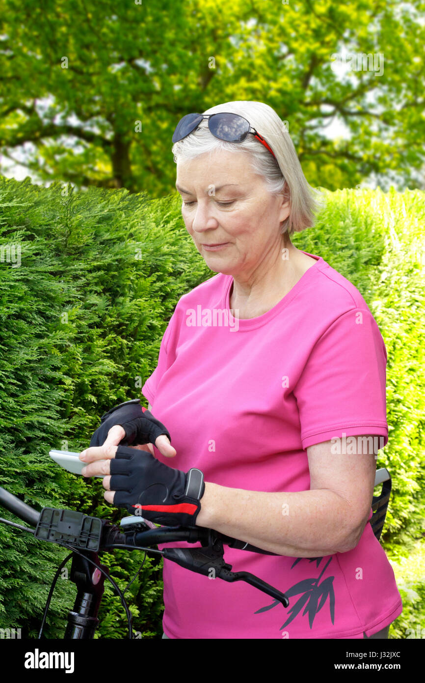 Ältere Frau im Freien im Sommer mit rosa Sport Shirt, Fahrrad Handschuhe und Sonnenbrille an Ihrem Smartphone, Hecke im Hintergrund suchen Stockfoto
