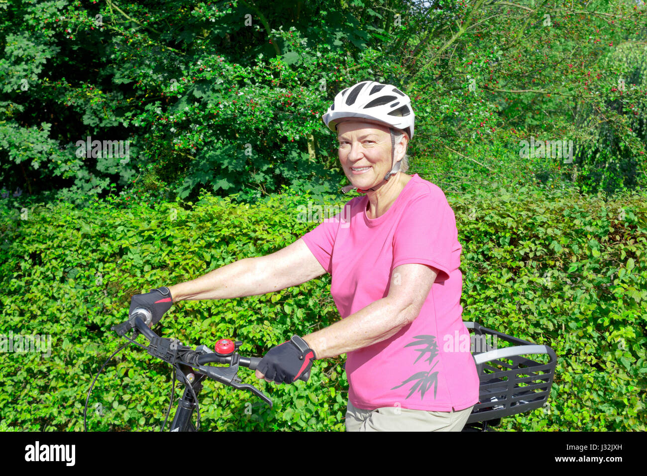 Lächelnde ältere Frau mit Helm und Handschuhe ihr Fahrrad Holding an einem sonnigen Tag, Hecke und Sträuchern im Hintergrund, kopieren oder Text Platz Stockfoto