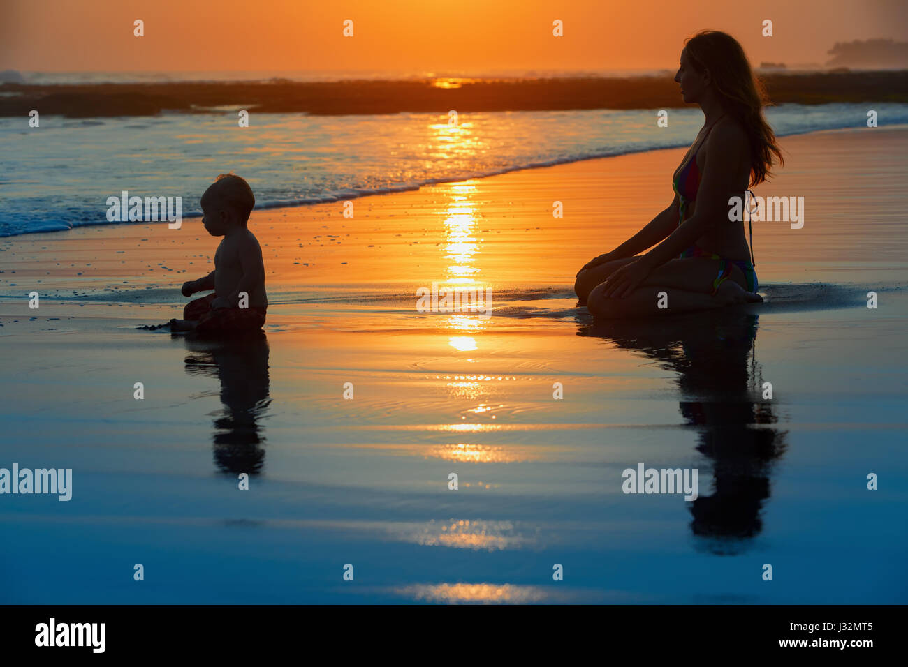 Familie Badespaß am schwarzen Sandstrand mit Meer Surfen. Glückliche Mutter, Baby Sohn sitzen und Ozean auf Sonnenuntergang Himmel mit Sonne Hintergrund betrachten. Kind mit pare Stockfoto