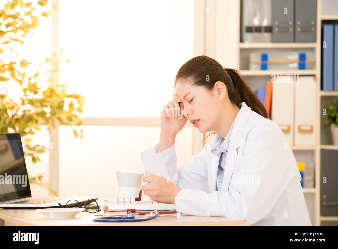 junge Ärztin fühlen sich müde und Kopfschmerzen in Klinik Büro Kaffee trinken. Gemischte Rassen asiatische chinesische Modell. Ärzte und medizinisches Konzept. Stockfoto