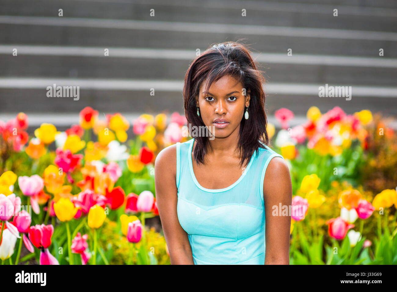 Young African American Frau trägt grüne Tank-Top, Ohrringe, auf Garten mit bunten Blumen in New York sitzen, relaxen, denken Stockfoto