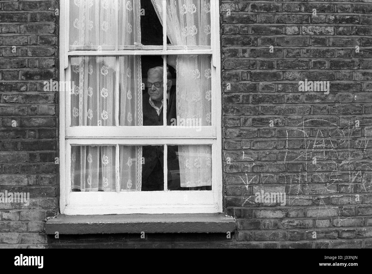 Allein ein älterer Mann, der aus dem Fenster blickt, durch Netzvorhänge 1970er Whitechapel, East End London 1975 UK HOMER SYKES Stockfoto