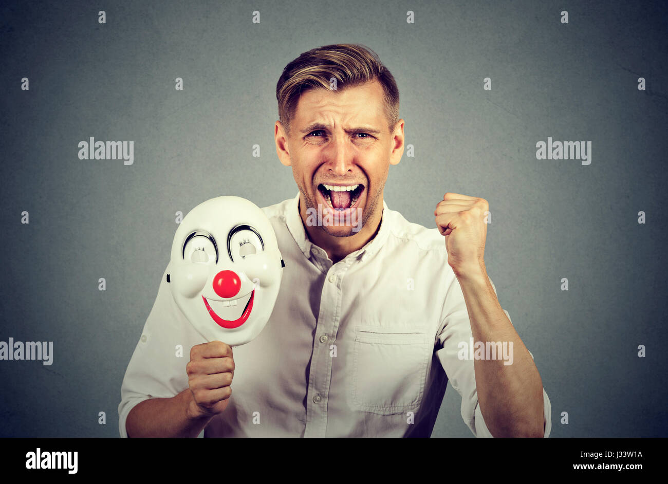 Porträt-junge verärgert böse schreienden Mann hält eine Clownsmaske mit dem Ausdruck ihrer Fröhlichkeit Glück auf graue Wand Hintergrund isoliert. Menschliche Emotionen ja Stockfoto