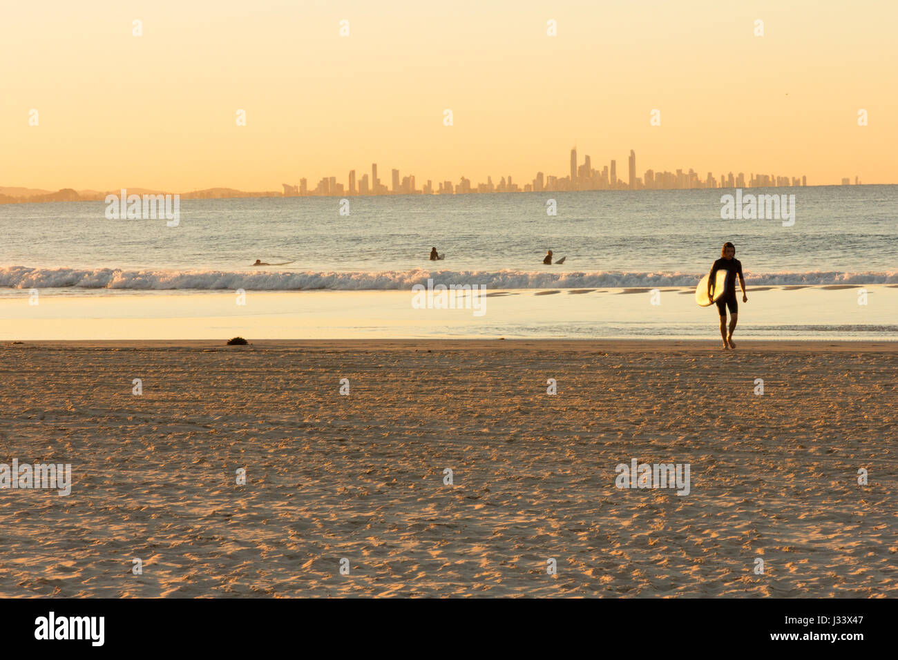 Surfer geht weg vom Meer auf Coolangatta Strand bei Sonnenuntergang mit Surfer's Paradise im Hintergrund Stockfoto