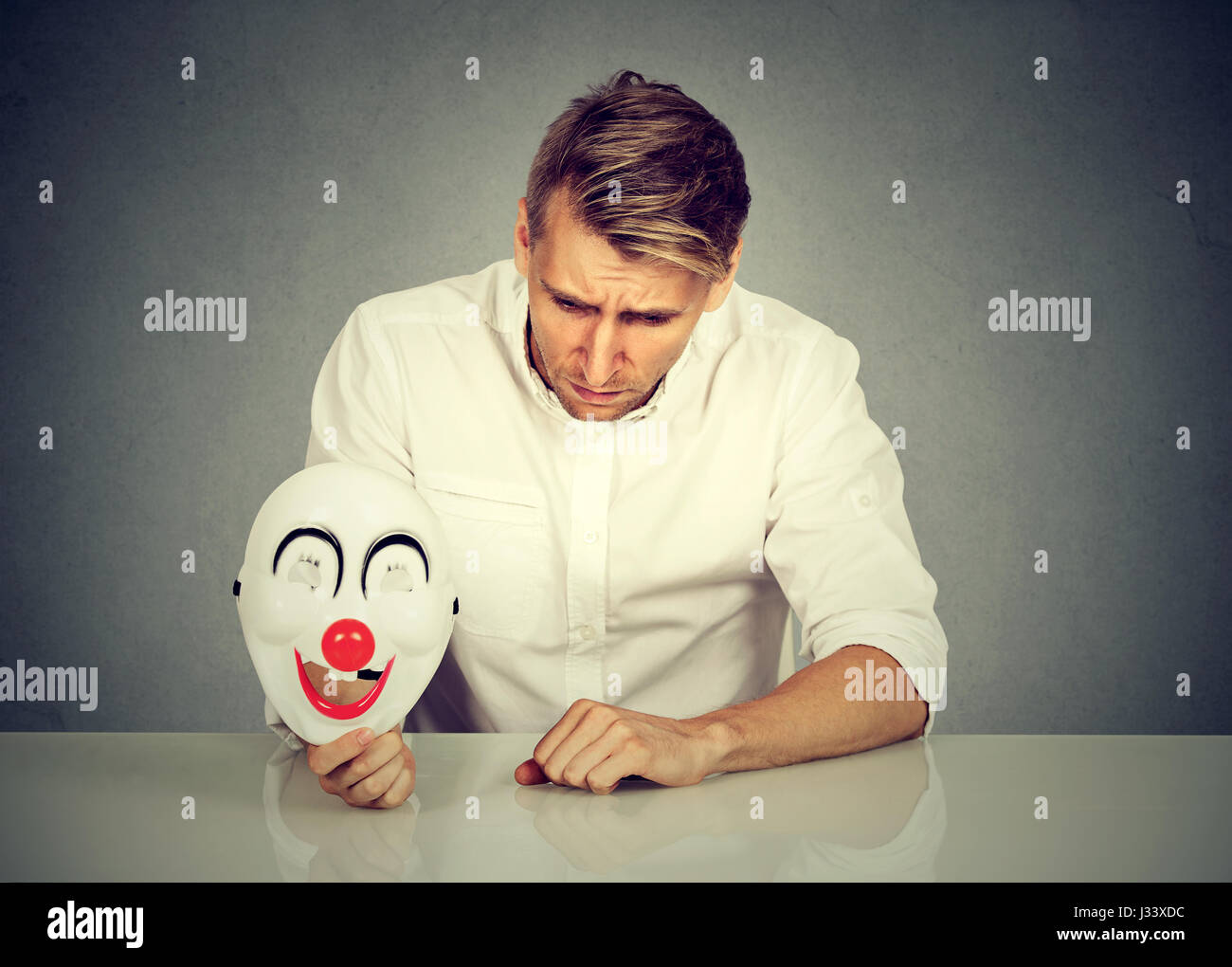 Porträt junge aufgeregt besorgt Mann mit traurigen Ausdruck Clownsmaske mit dem Ausdruck ihrer Fröhlichkeit Glück auf graue Wand Hintergrund isoliert halten. Menschlichen emot Stockfoto