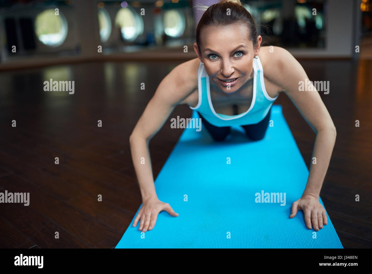 Blonde Frau mittleren Alters Liegestütze trainieren im Fitness-Studio und Blick in die Kamera beim liegen auf blaue Yoga Teppich über Spiegel Hintergrund Stockfoto