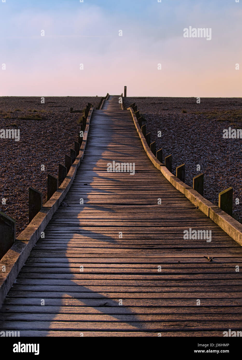 Einen schmalen Holzsteg erstreckt sich von der Kamera zum Horizont auf einem Kiesstrand in niedrigen gerichtetes Sonnenlicht mit blauen Himmel und Wolken über. Stockfoto