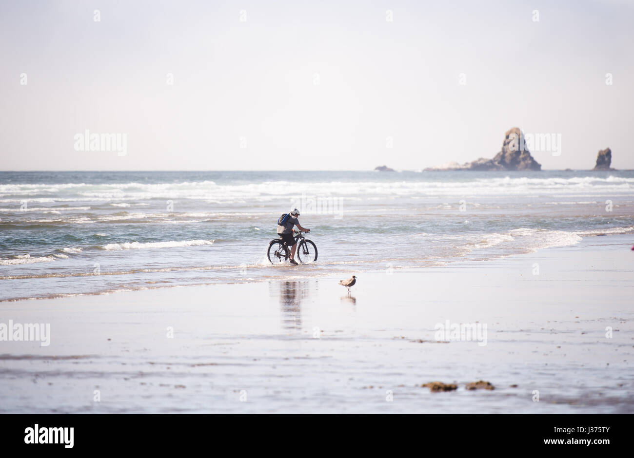 Ein kleiner Junge mit einem Rucksack auf den Schultern reitet ein Fahrrad auf dem Wasser unter den schäumenden Wellen an der Pazifikküste, erleben ihre eigene Stockfoto