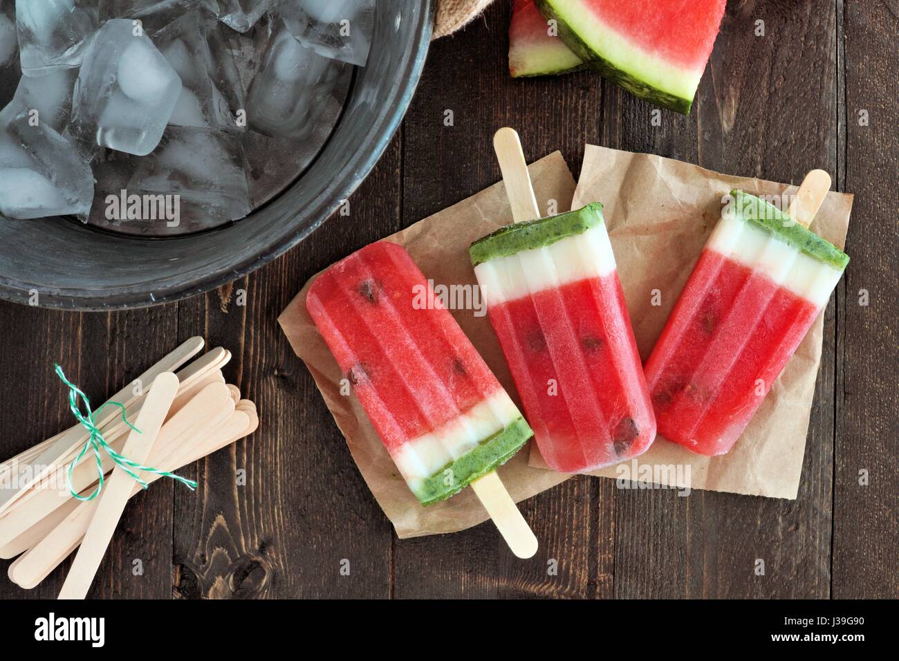 Hausgemachte Wassermelone Eis am Stiel auf Papier mit Eiskübel und rustikale Holz Hintergrund Stockfoto