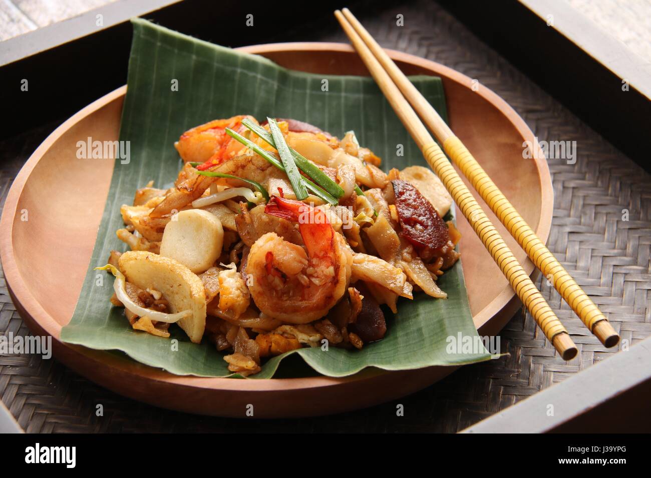 Char Kway Teow. Flache Reisnudeln mit Garnelen, Wurst, Sojabohnensprossen, Ei und Soja-Sauce gebraten. Auf einer hölzernen Platte ausgekleidet mit Bananenblatt serviert. Stockfoto