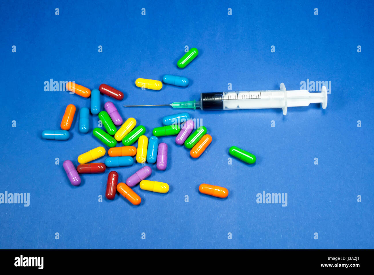 Medizinische Spritze teilweise gefüllt mit Flüssigkeit, horizontal platziert unter einem Haufen von verschiedenen farbigen Pillen, medizinisches Konzept, blauer Hintergrund Stockfoto