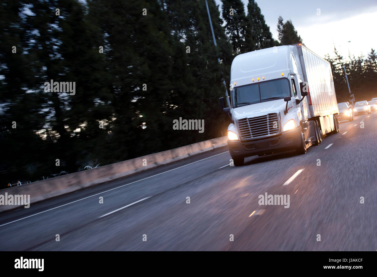 Eine moderne Big Rig Semi Truck mit Anhänger und Lichter auf die Bewegung entlang der Abend direct Highway ist mit einer Reflexion des Lichts der Scheinwerfer Stockfoto