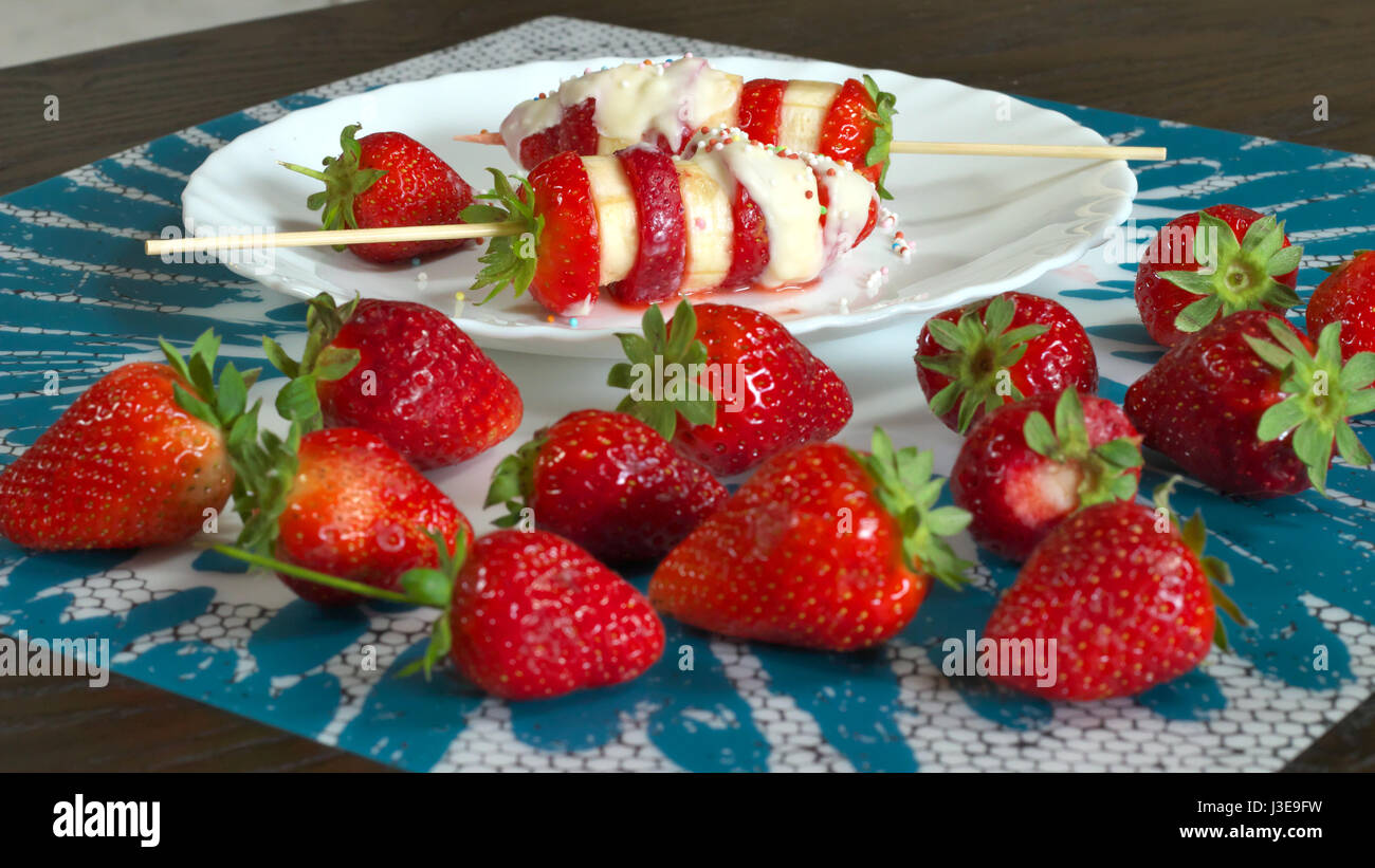 Erdbeere und Banane Spieße vorbereitet Rezept mit weißer Schokolade und bunten Zucker Dekorationen - rote Erdbeeren im Vordergrund servierbereit Stockfoto