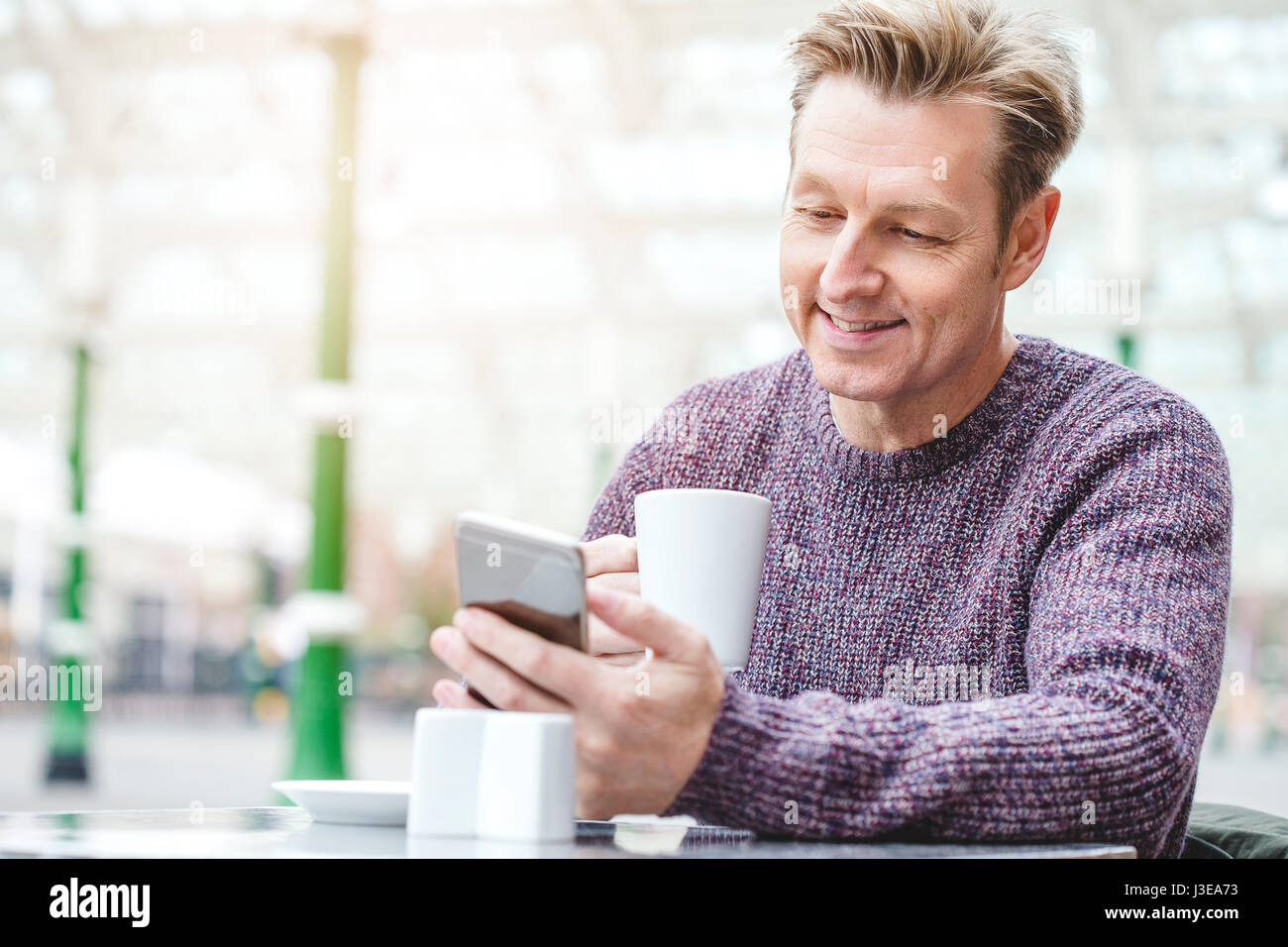 Mann bei einem heißen Getränk in einem Café in einem Bahnhof. Er sitzt außerhalb, blickte auf sein Smartphone in der Hand ist. Stockfoto