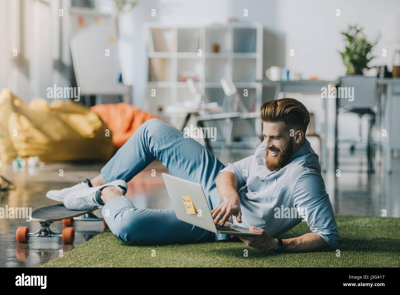 Bärtigen Mann am Boden liegend und mit Laptop in Kreativbüro lächelnd Stockfoto