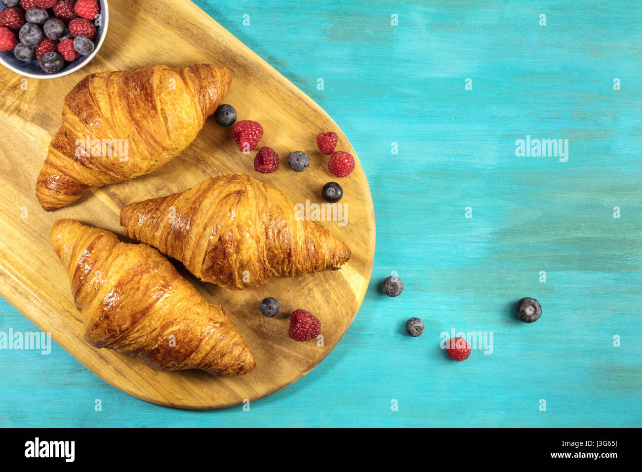 Eine obenliegende Aufnahme des knackigen französische Croissants mit frischen Himbeeren und Heidelbeeren auf einem Holzbrett und lebhaften Türkis Textur mit cop Stockfoto