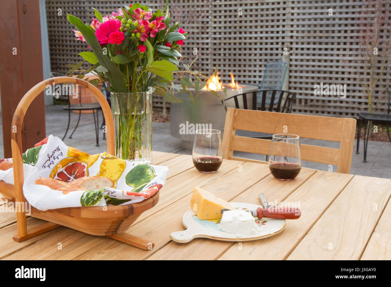 Zwei Gläser Rotwein auf Holztisch mit Picknick-Korb und Keil von Käse auf Teller mit Messer. Blumen am Tisch, Feuerstelle mit Feuer im Hintergrund. Stockfoto