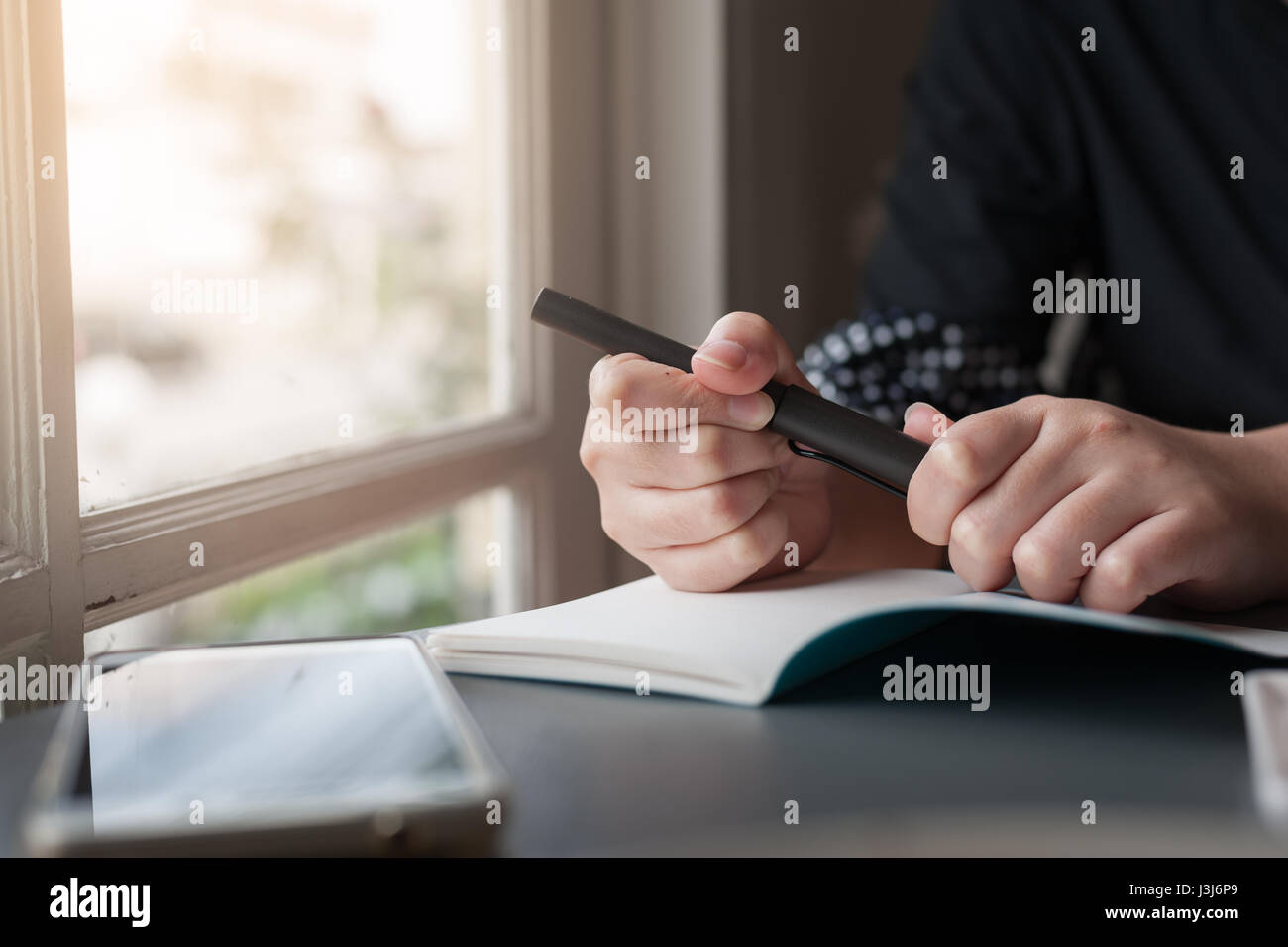Frau Hand mit Stift beim Schreiben auf kleinen Notebook neben Fenster. Freier Journalist in Heimarbeit Konzept. Stockfoto