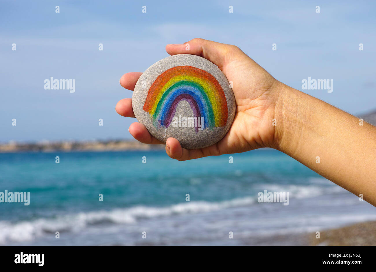 Frau Hand halten Kiesel mit bemalten Regenbogen gegen blauen Himmel und Meer. Stockfoto