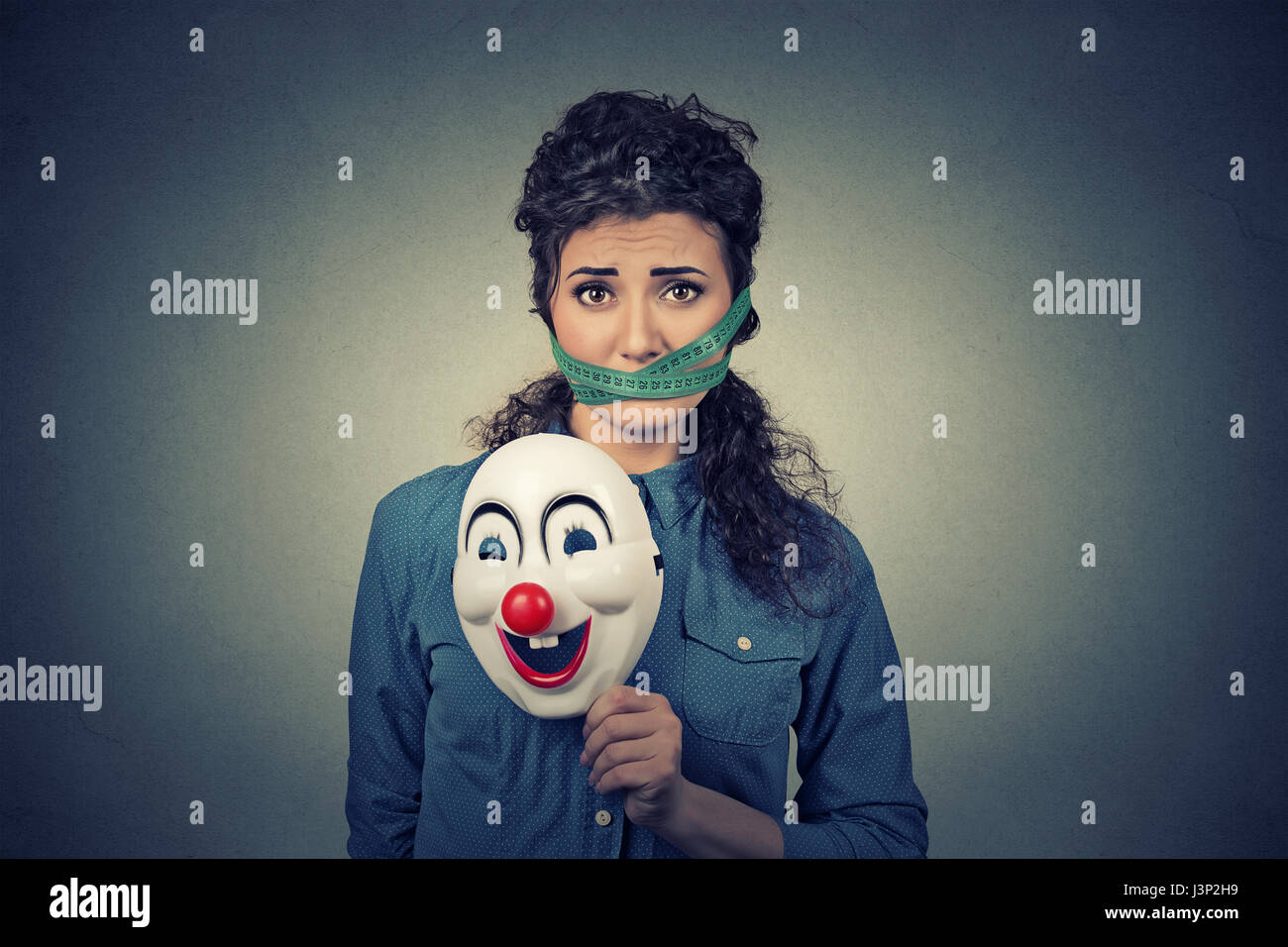 Diät-Einschränkung und Stress-Konzept. Porträt des jungen traurige Frau mit Clownsmaske und Maßband um den Mund auf graue Wand Hintergrund isoliert. Stockfoto