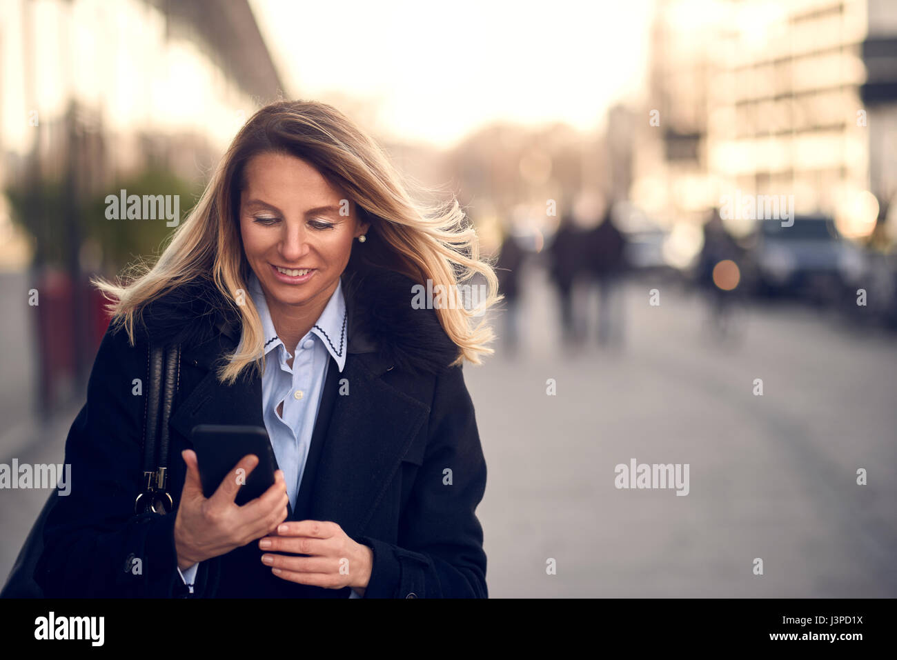 Modische Frau mittleren Alters im schwarzen Mantel und blonden Haaren beschäftigt mit ihrem Mobiltelefon während einer Stadtstraße Stockfoto