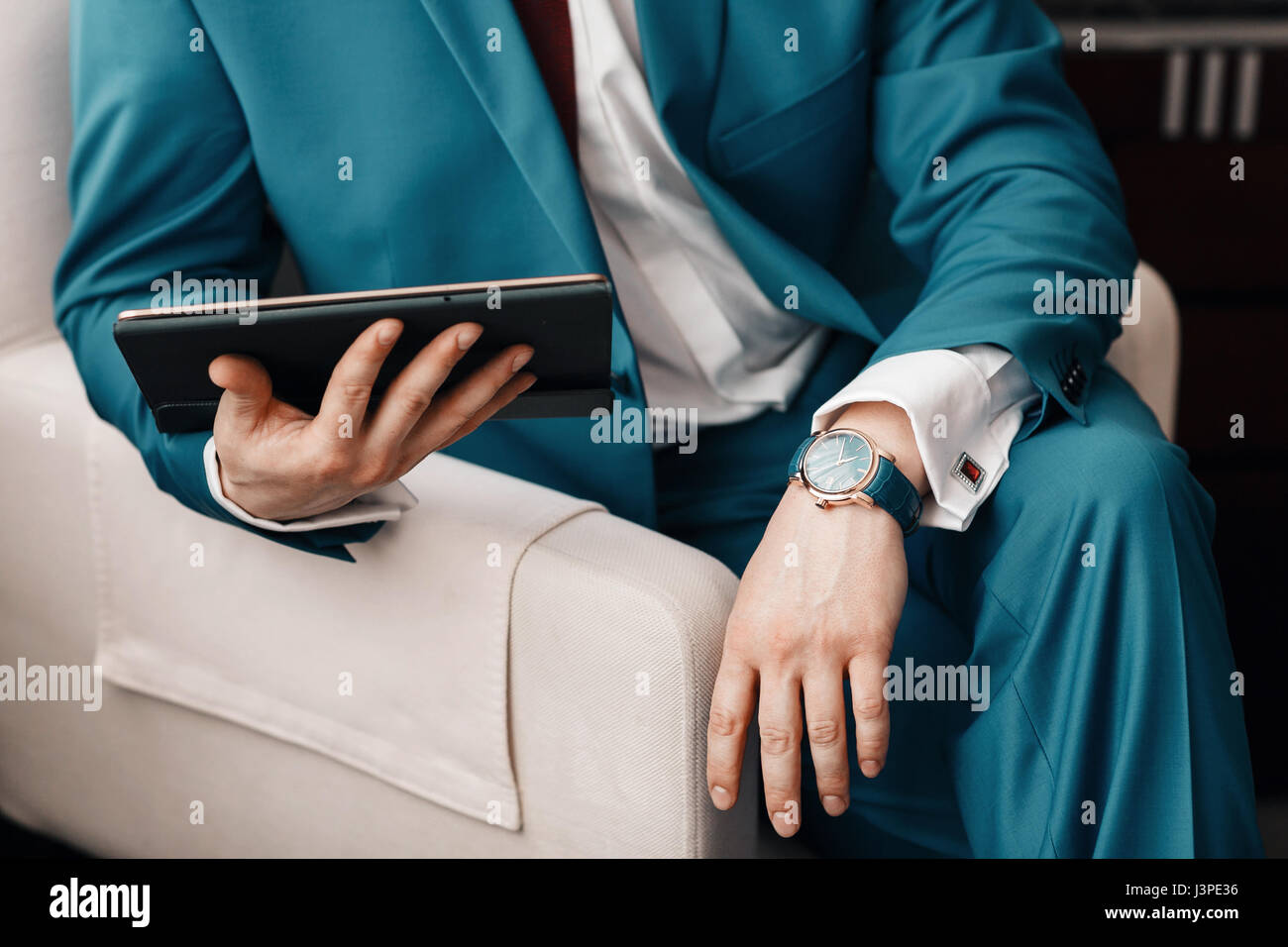 Geschäftsmann halten Sie ein digitale Tablet in der hand beim Sitzen auf einem Sofa in einem blauen Anzug. auf Hand teure mechanische Uhr mit Lederarmband. Hemd mit Manschettenknöpfen Stockfoto