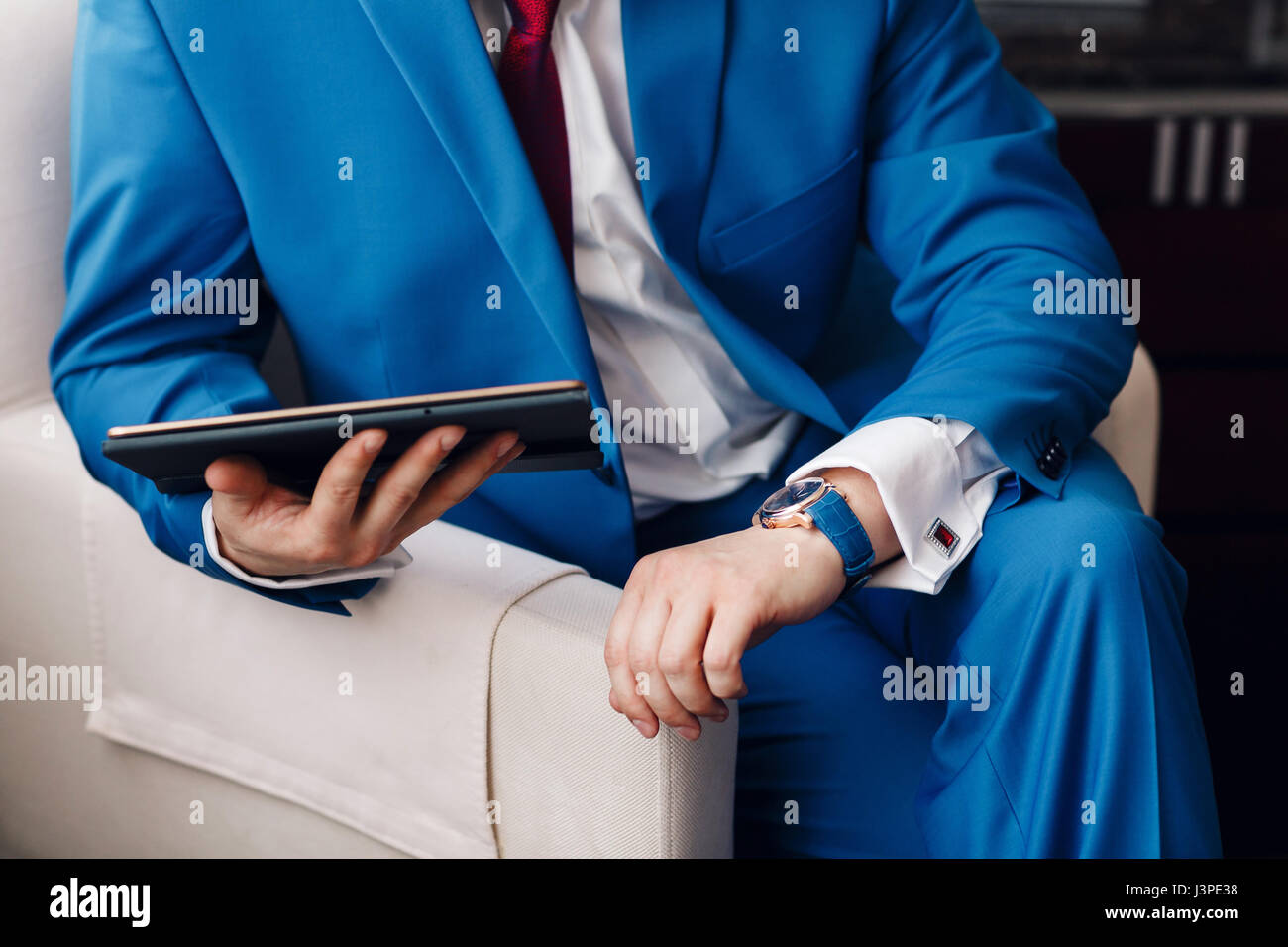 Geschäftsmann halten Sie ein digitale Tablet in der hand beim Sitzen auf einem Sofa in einem blauen Anzug. auf Hand teure mechanische Uhr mit Lederarmband. Hemd mit Manschettenknöpfen Stockfoto
