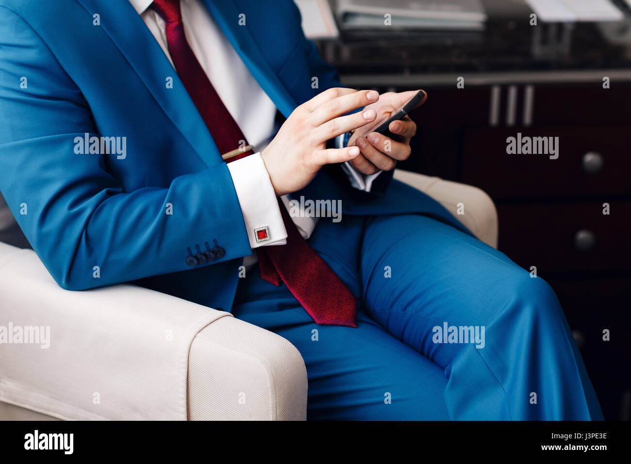 Geschäftsmann halten Sie eine Telefon in der hand beim Sitzen auf einem Sofa in einem blauen Anzug. auf Hand teure mechanische Uhr mit Lederarmband. Hemd mit Manschettenknöpfen Stockfoto