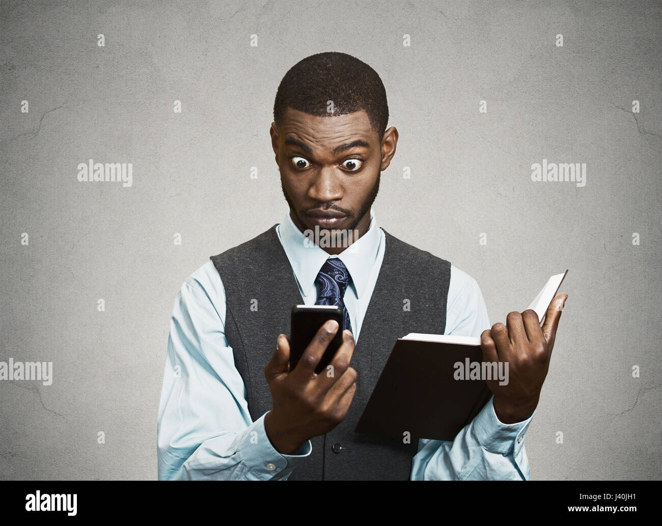 Closeup Portrait überrascht, schockiert Business Mann executive Lesung breaking News auf Smartphone hält Buch isoliert schwarz grau hinterlegt. Menschliches Gesicht Stockfoto