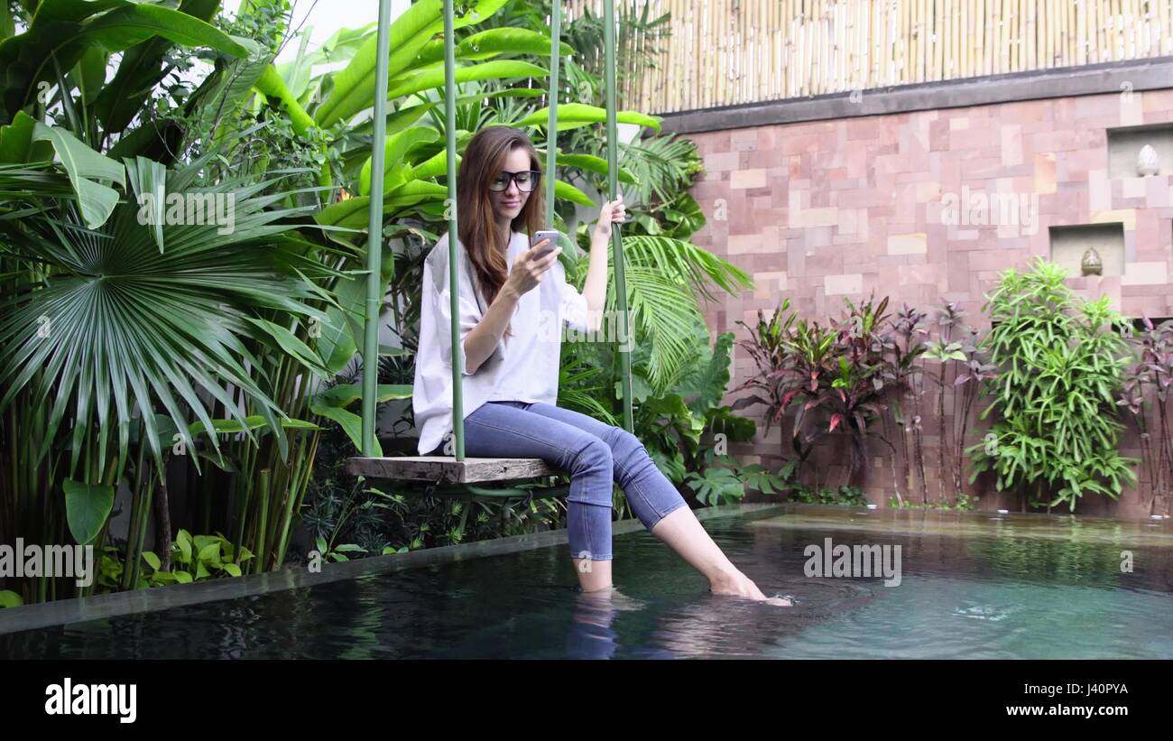 Junge Frau in Gläsern mit Smartphone sitzend auf Schaukel in der Nähe von  Pool im tropischen Land. 4K Stockfotografie - Alamy