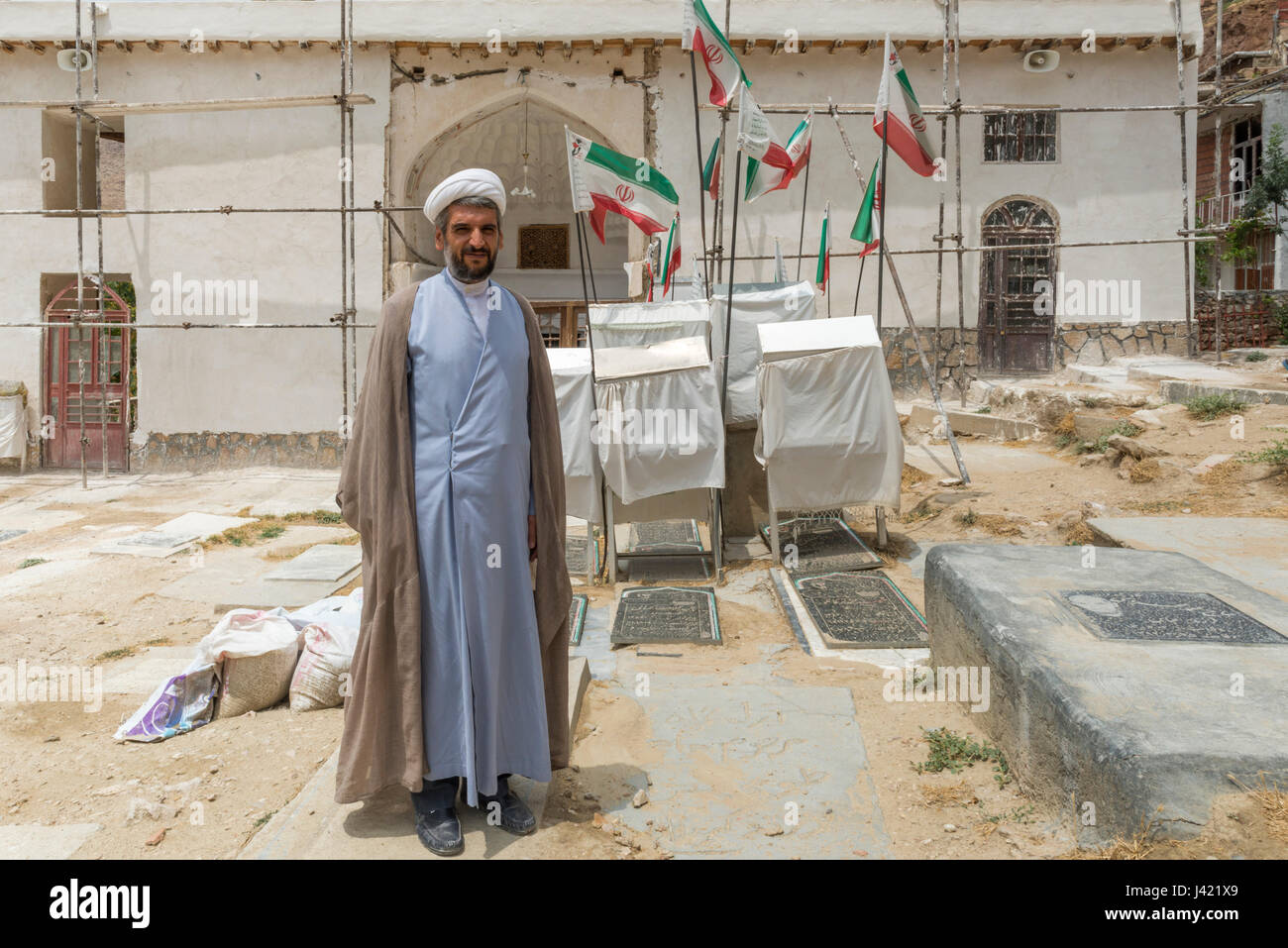 Akhoond Kleriker (oder Mullah) auf dem Friedhof mit iranischen Fahnen vor der örtlichen Moschee, Esfidan, Nord-Chorasan, Iran Stockfoto