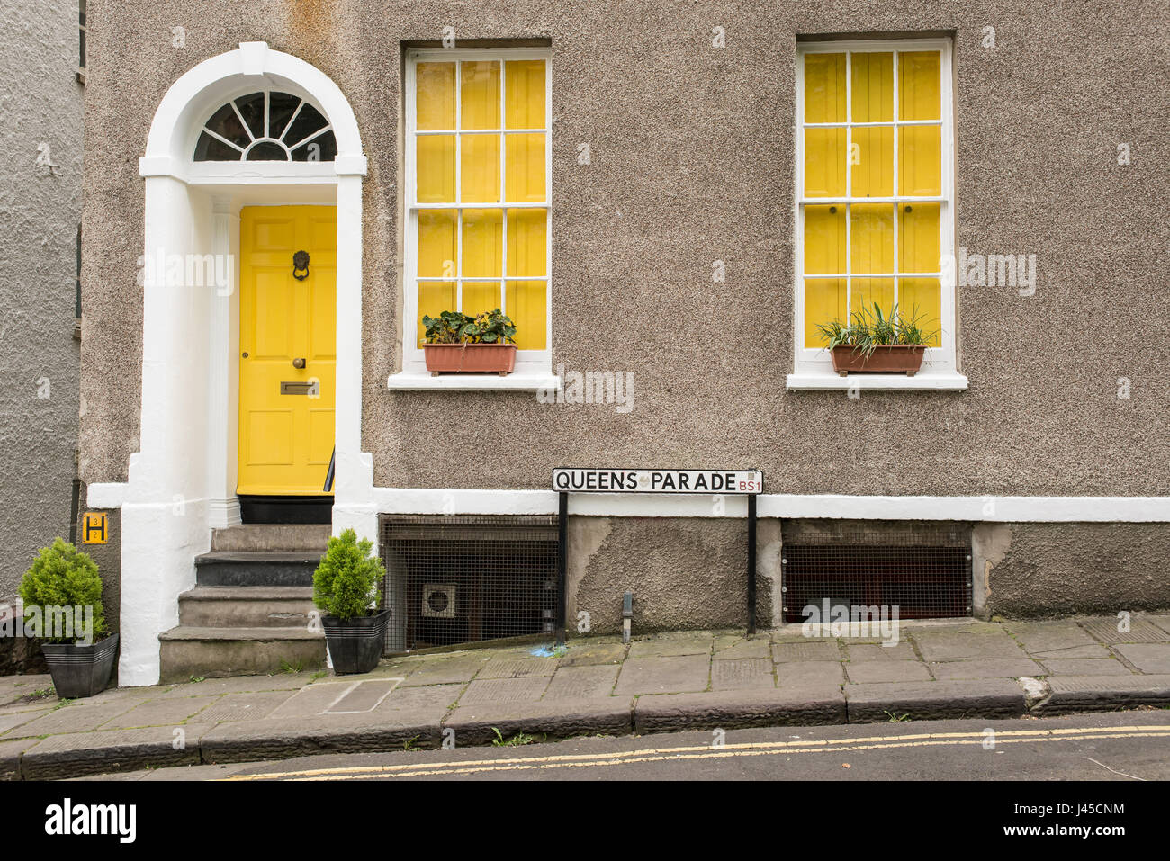 Exterieur des britischen Traditionshauses in Queens Parade, Bristol, UK. Mit grauen Wänden, gelbe vordere Tür und zwei Fenstern mit gelben Vorhänge. Blumen Stockfoto