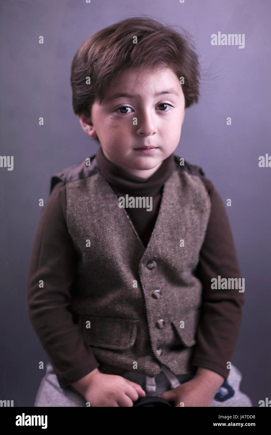 Porträt einer hübschen kleinen Jungen mit einem braunen rollkragen pullover und Brown tweed Weste mit Kopf voll dickes braunes Haar vor grauem Hintergrund Stockfoto