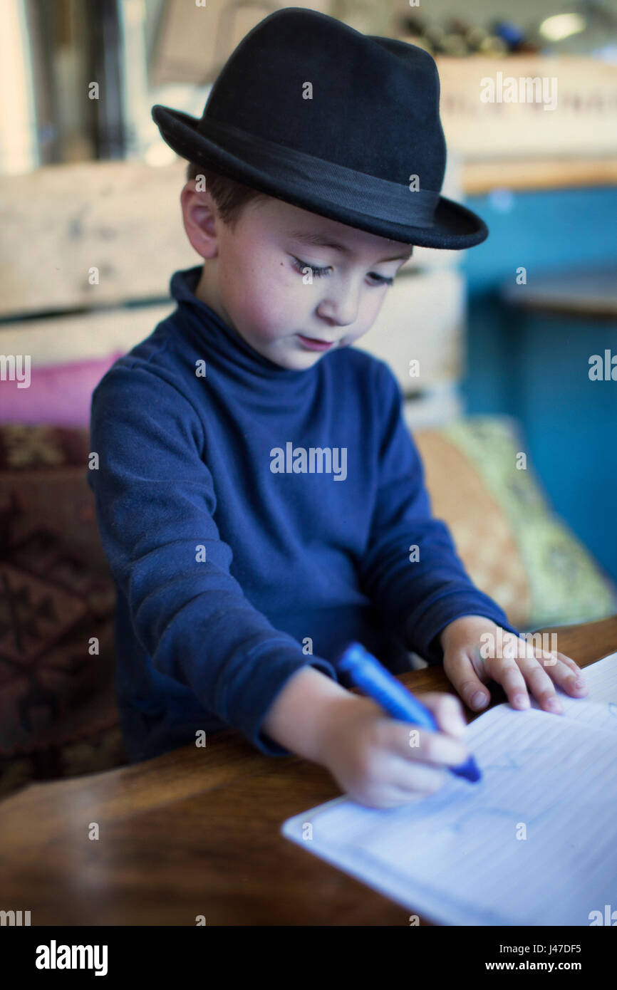 Süße kleine Junge mit braunen Haaren trägt einen schwarzen fedora Hut und ein blauer rollkragen pullover Färbung in einem Buch mit einem blauen crayola Crayon Stockfoto