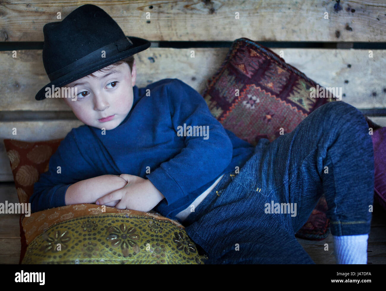 Süße kleine Junge mit braunen Haaren trägt einen schwarzen fedora Hut und ein blauer rollkragen pullover liegend auf einem hölzernen Sitzbank mit Kissen Stockfoto
