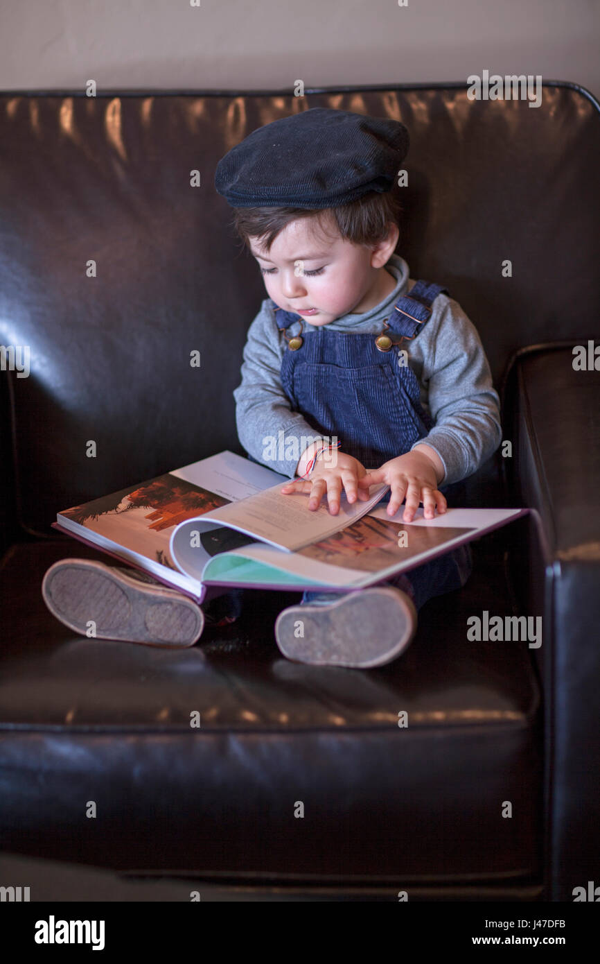 Süße kleine Junge mit braunen Haaren trägt ein blaues Denim Jeans Overalls und einem blauen Cord Cap hat ein Buch lesen in einem braunen Leder Stuhl Stockfoto