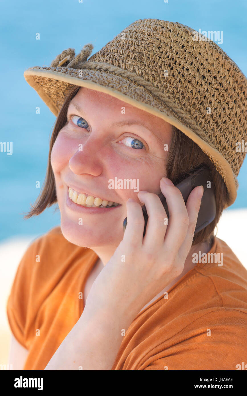 Schöne Erwachsene kaukasischen Frau tragen stilvolle Strohhut ist Mobiltelefon in den Sommerferien Urlaub am Meer Stockfoto