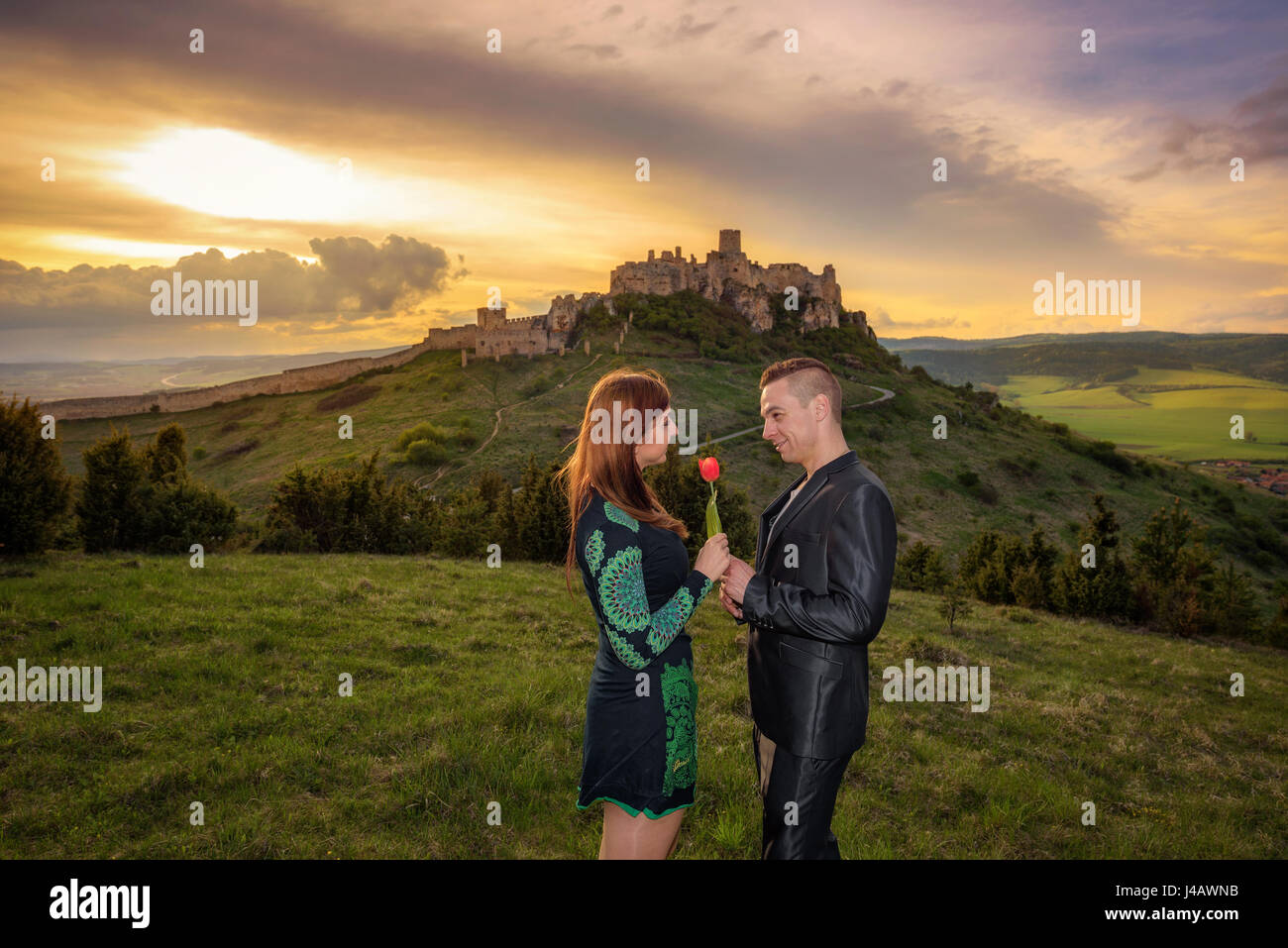 Junges Paar in Liebe bei Sonnenuntergang in der Nähe einer Burgruine. Der Mann gibt eine rote Tulpe zu seiner Freundin. Stockfoto