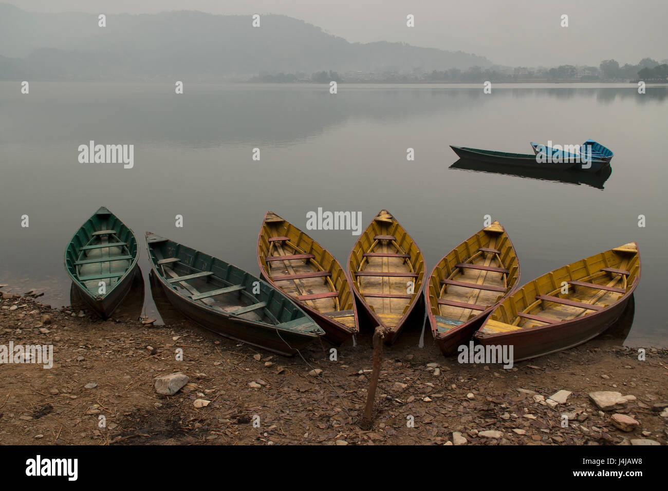 Grüne und gelbe Holzboote am Ufer des Sees Feva, das ruhige Wasser des Sees als Spiegel reflektiert den Blue Mountains im Hintergrund, Pokhara Stockfoto