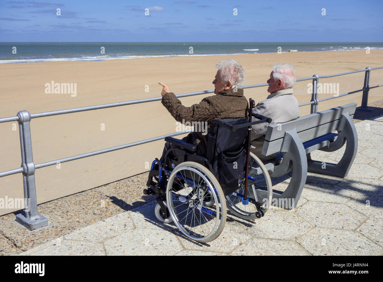 Ältere Frau im Rollstuhl und pensionierte Mann sitzt auf einer Bank am Promenade entlang der Küste an einem kalten, sonnigen Tag im Frühjahr deaktiviert Stockfoto