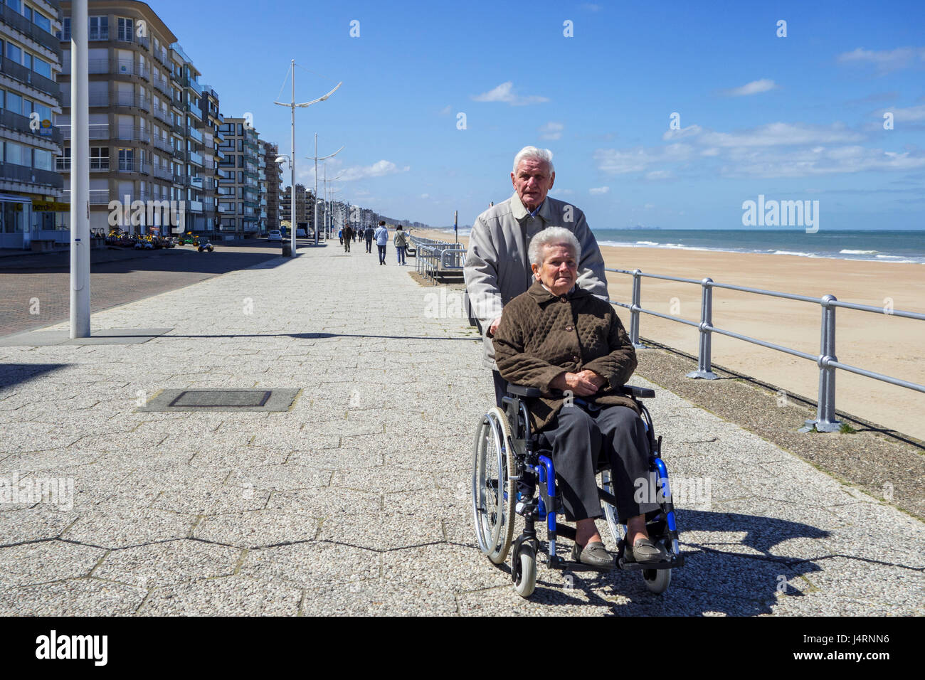 Pensionierter Mann unter deaktiviert ältere Frau im Rollstuhl für einen Spaziergang auf der Promenade entlang der belgischen Nordseeküste an einem kalten, sonnigen Tag im Frühling Stockfoto