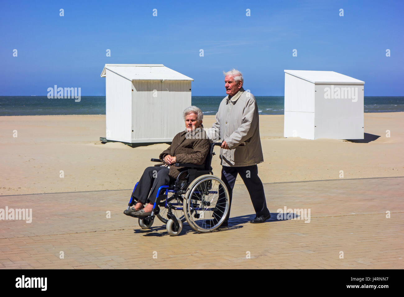 Pensionierter Mann unter deaktiviert ältere Frau im Rollstuhl für einen Spaziergang auf der Promenade entlang der Küste an einem kalten, sonnigen Tag im Frühling Stockfoto