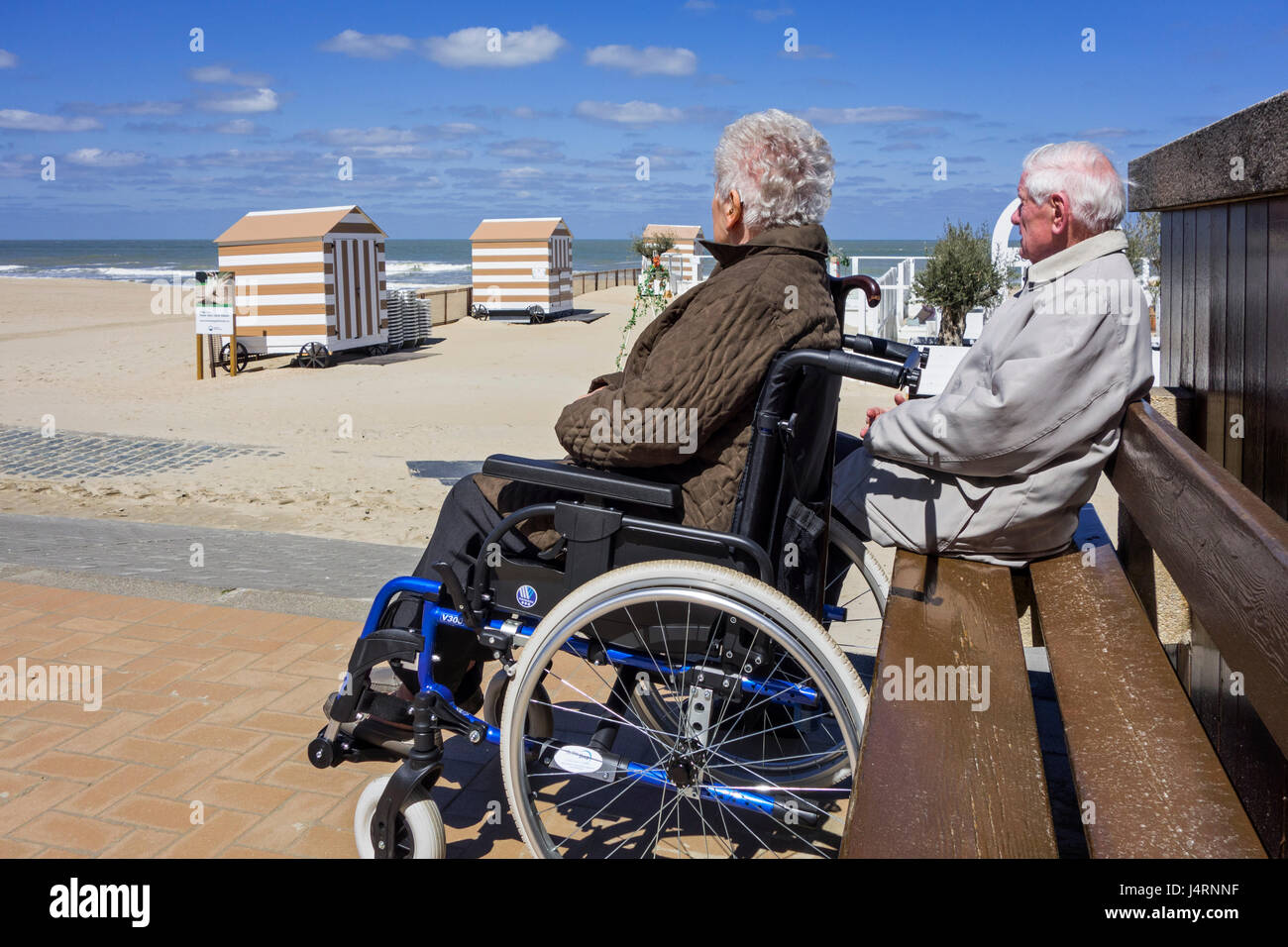 Ältere Frau im Rollstuhl und pensionierte Mann sitzt auf einer Bank am Promenade entlang der Küste an einem kalten, sonnigen Tag im Frühjahr deaktiviert Stockfoto