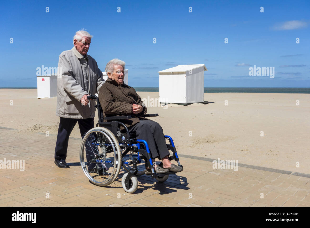Pensionierter Mann unter deaktiviert ältere Frau im Rollstuhl für einen Spaziergang auf der Promenade entlang der Küste an einem kalten, sonnigen Tag im Frühling Stockfoto