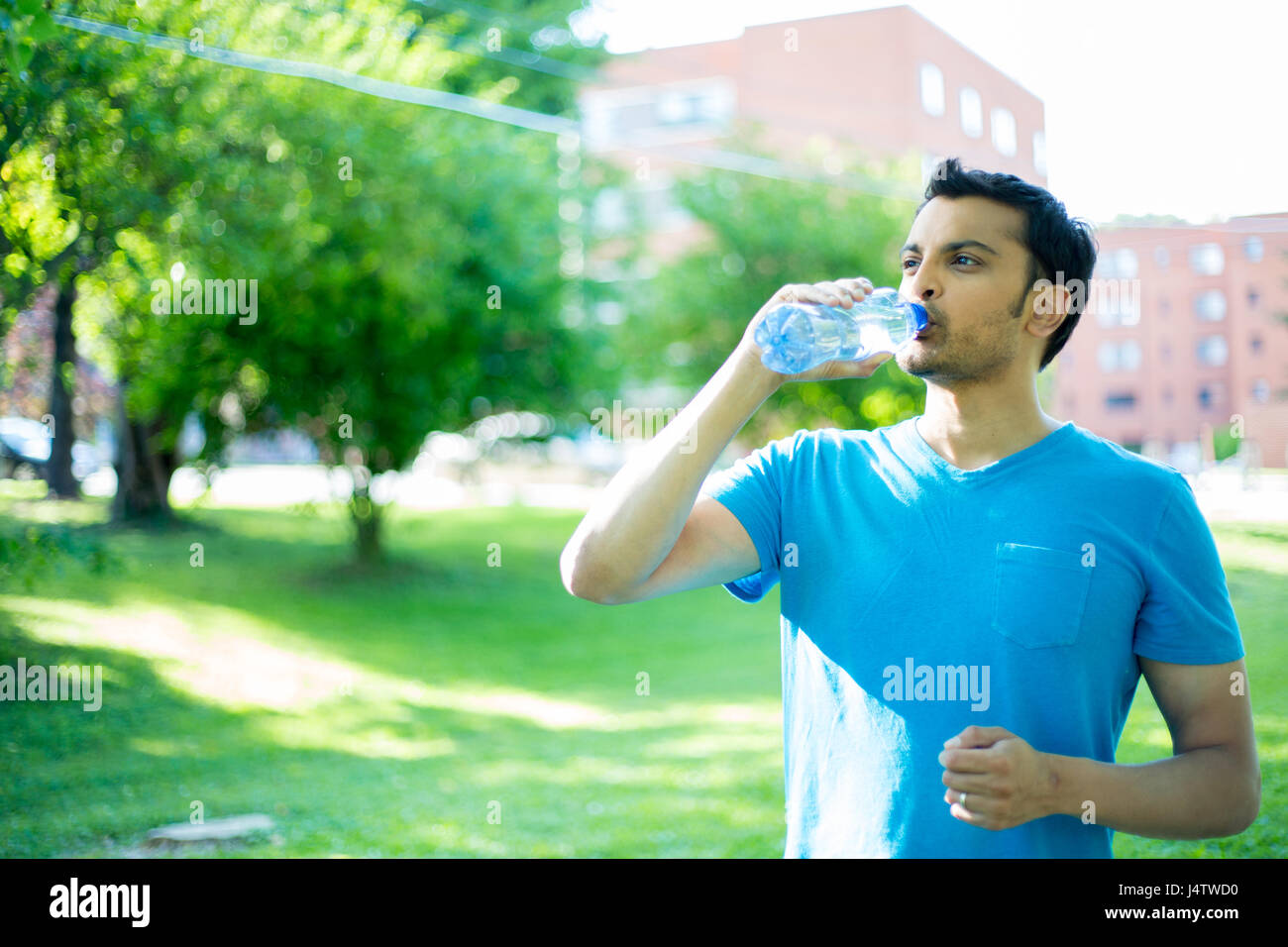 Closeup Portrait der junge Mann im blauen Hemd Trinkwasser aus kristallklarem Flasche auf einem heißen, sonnigen Tag, isolierten Grün der Bäume und Gebäude-Hintergrund Stockfoto