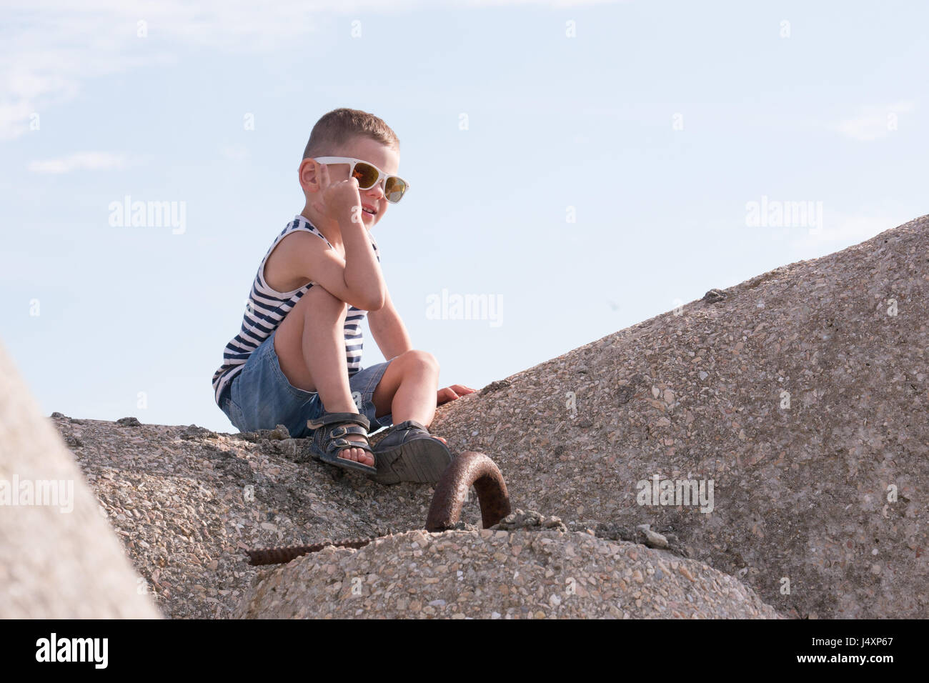 fröhliche kleine Junge in Sonnenbrillen, Sailor Streifen Weste und Shorts sitzen auf einem Wellenbrecher lächelnd Stockfoto