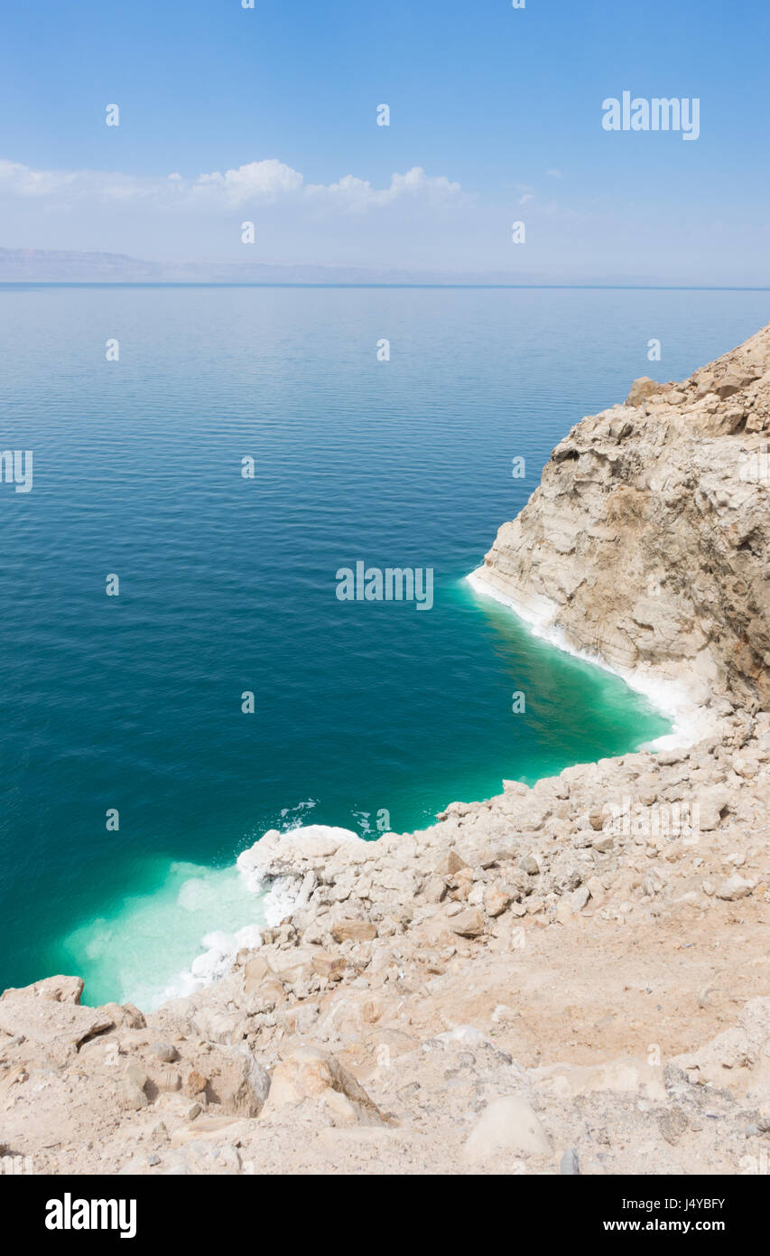 Mit Blick auf das Tote Meer mit Türkis und Aquamarin Wasser und robust, Salz verkrusteten Bank. Der Horizont ist diesig mit blauen Himmel und Wolken. Stockfoto